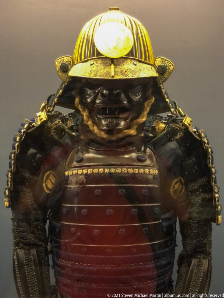 Close frontal view of Japanese samurai armor with striped helmet, dark face guard, layered shoulders, and red-brown cuirass.