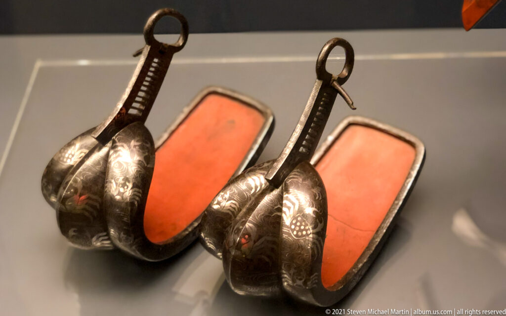 Pair of dark metal stirrups with tall suspension loops, engraved decoration, and bright orange-red footbeds.