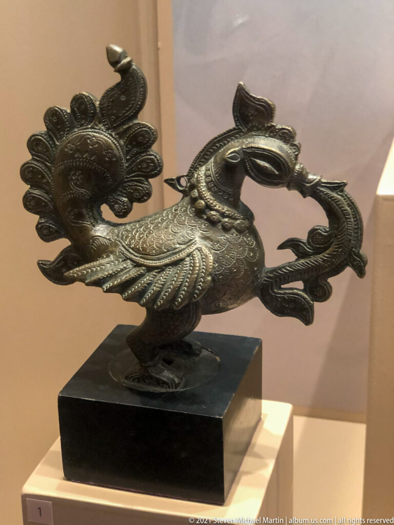 Indian bronze bird with raised patterned wings, high crest, curved neck, and elaborate scrolling tail, dated 17th–19th century.