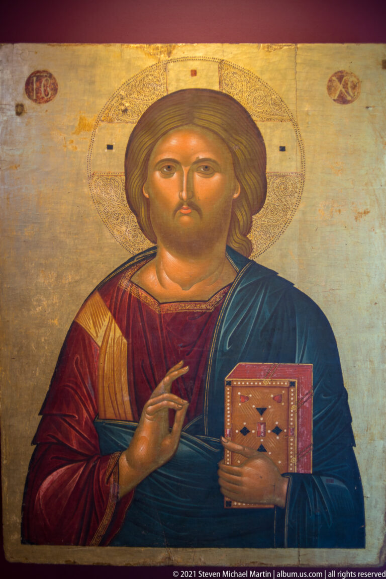 Bust-length Christ Pantocrator with cruciform halo, blessing hand, red and blue robes, and decorated Gospel book.