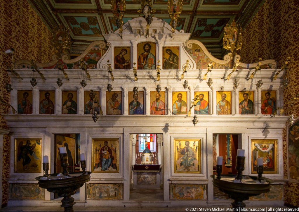 White carved iconostasis with rows of saint icons, larger lower panels, hanging lamps, and dark church furnishings.
