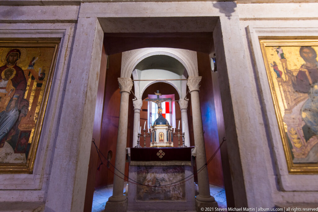 Small church altar framed by pale columns and an arch, with candles, a tabernacle-like object, stained glass, and flanking icons.