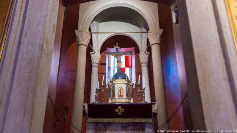 Close view of arched side altar with slender stone columns, candles, a small metal shrine, and colored window panes behind.