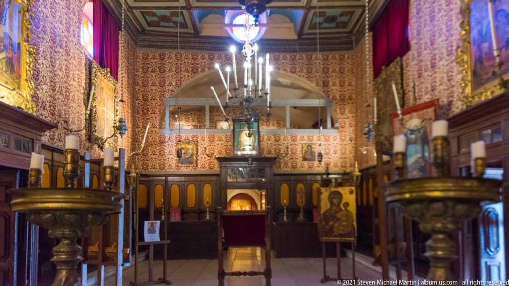 Interior of Antivouniotissa with dark wooden stalls, red patterned walls, chandeliers, brass candle stands, and devotional images.