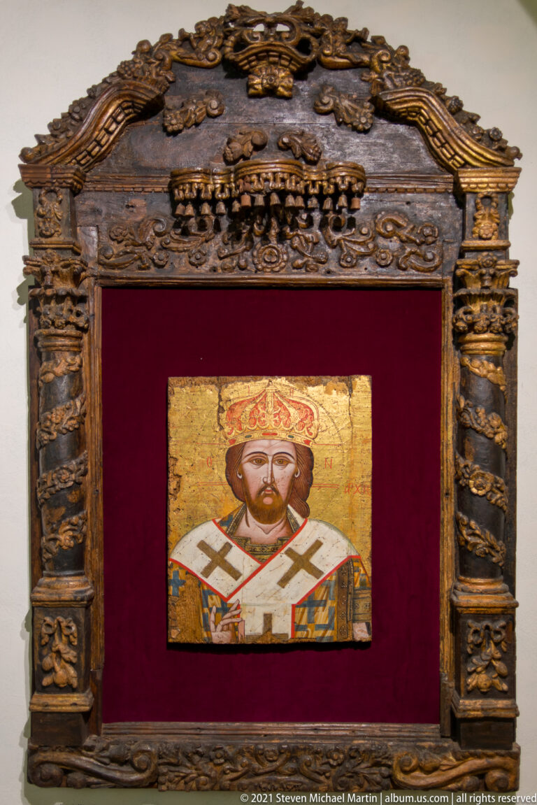 Small gold-ground icon of a crowned Orthodox hierarch in a large carved wooden frame with red backing.