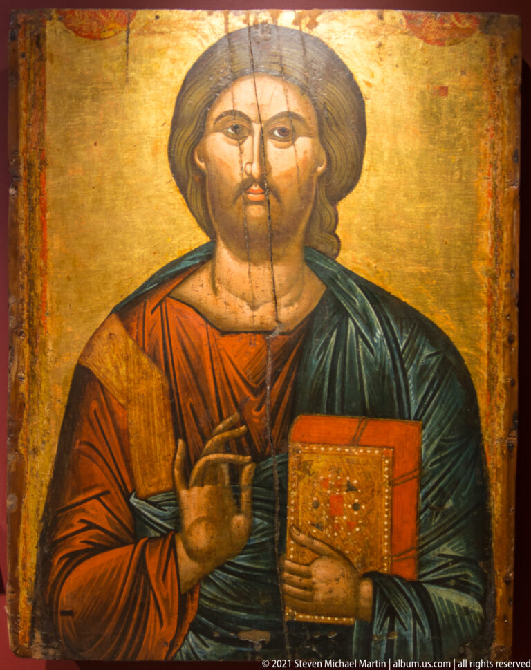 Close bust icon of Christ Pantocrator blessing and holding a decorated red Gospel book against a gold ground.