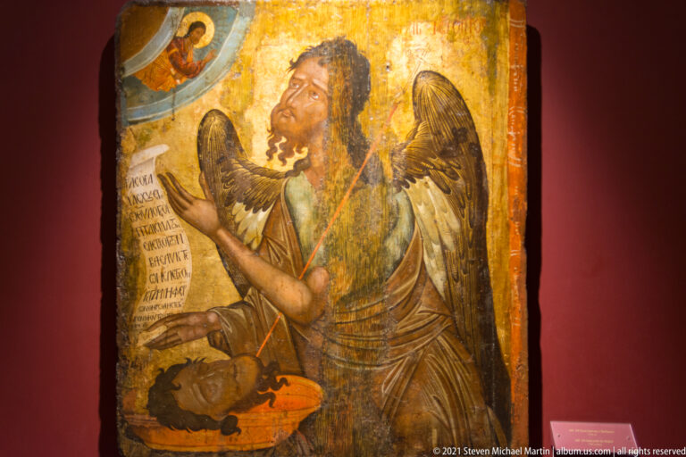 Winged Saint John the Baptist looks upward holding an inscribed scroll, with his severed head on a platter below.
