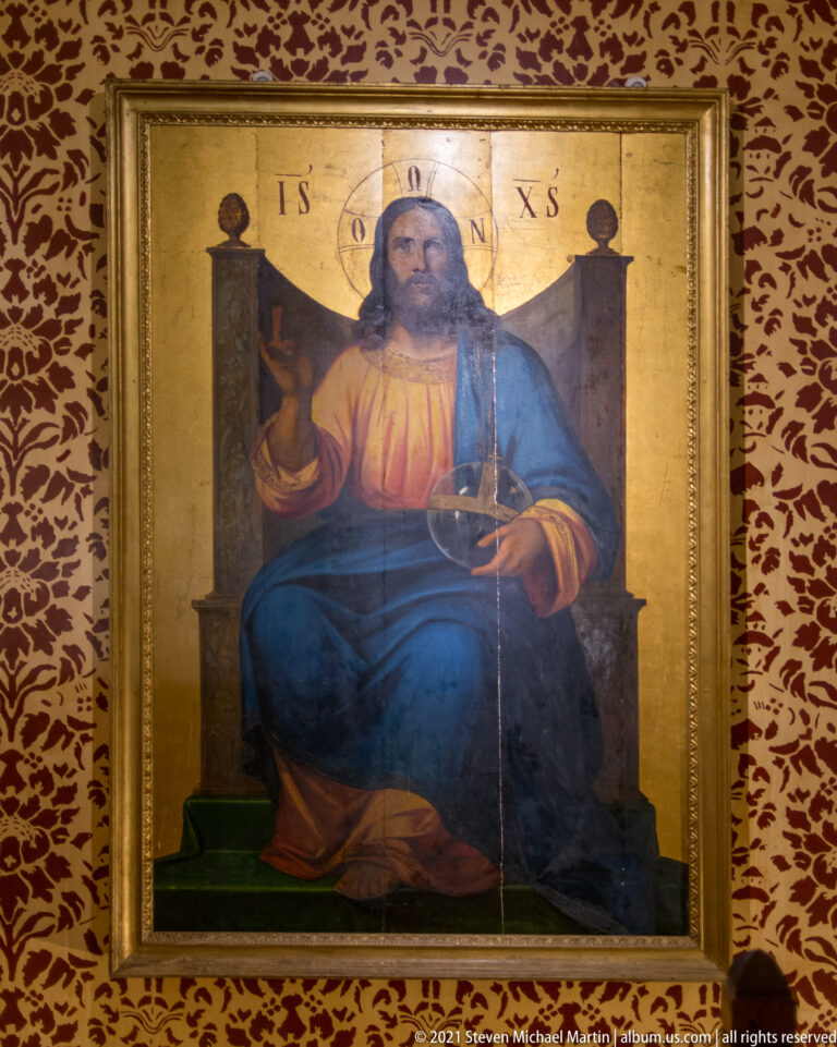 Framed icon of Christ seated frontally on a throne against a gold ground, displayed on a red patterned wall.