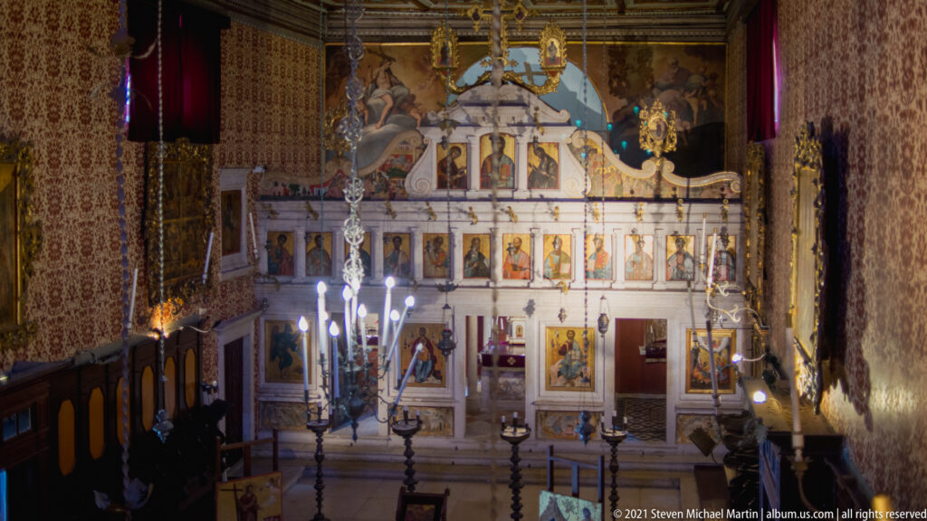 Elevated interior view toward the tall white iconostasis, with rows of icons, chandeliers, and candle stands in the foreground.
