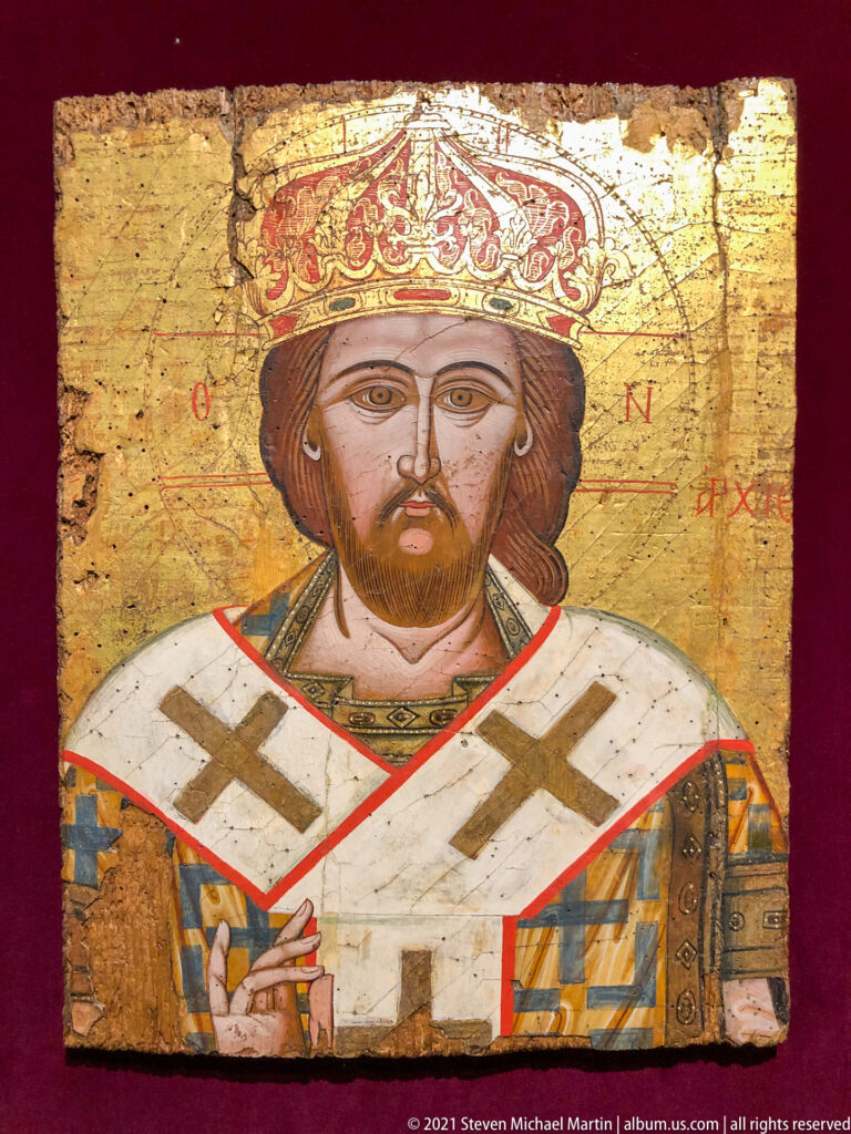 Bust icon of a crowned bearded Orthodox bishop wearing a cross-marked omophorion, blessing and holding a Gospel book.