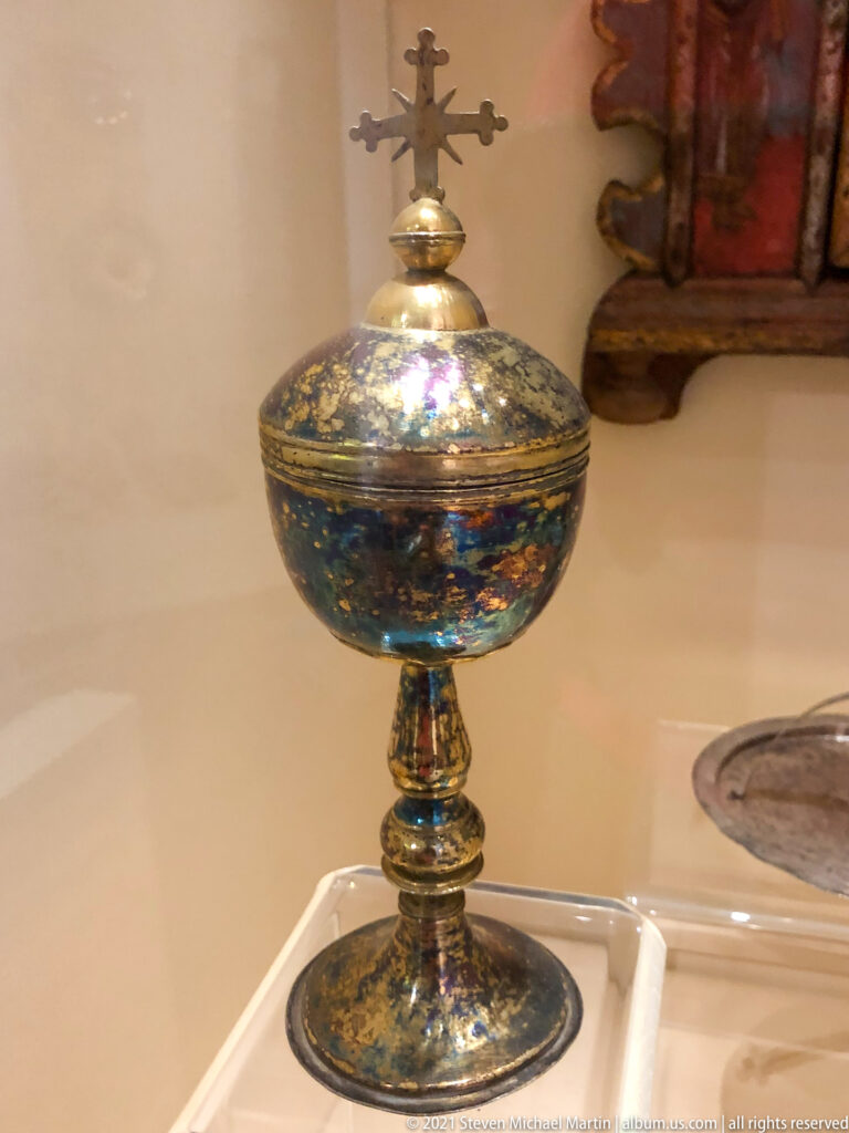 Small dark blue and gilded footed liturgical vessel with domed lid and cross finial in a museum case.