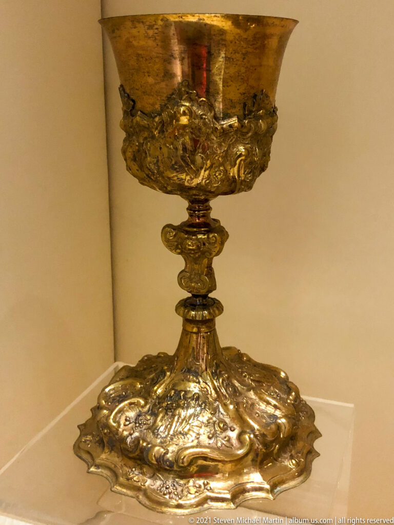 Gilded liturgical chalice with broad cup, ornate knopped stem, and lobed foot covered in raised scrolling decoration.