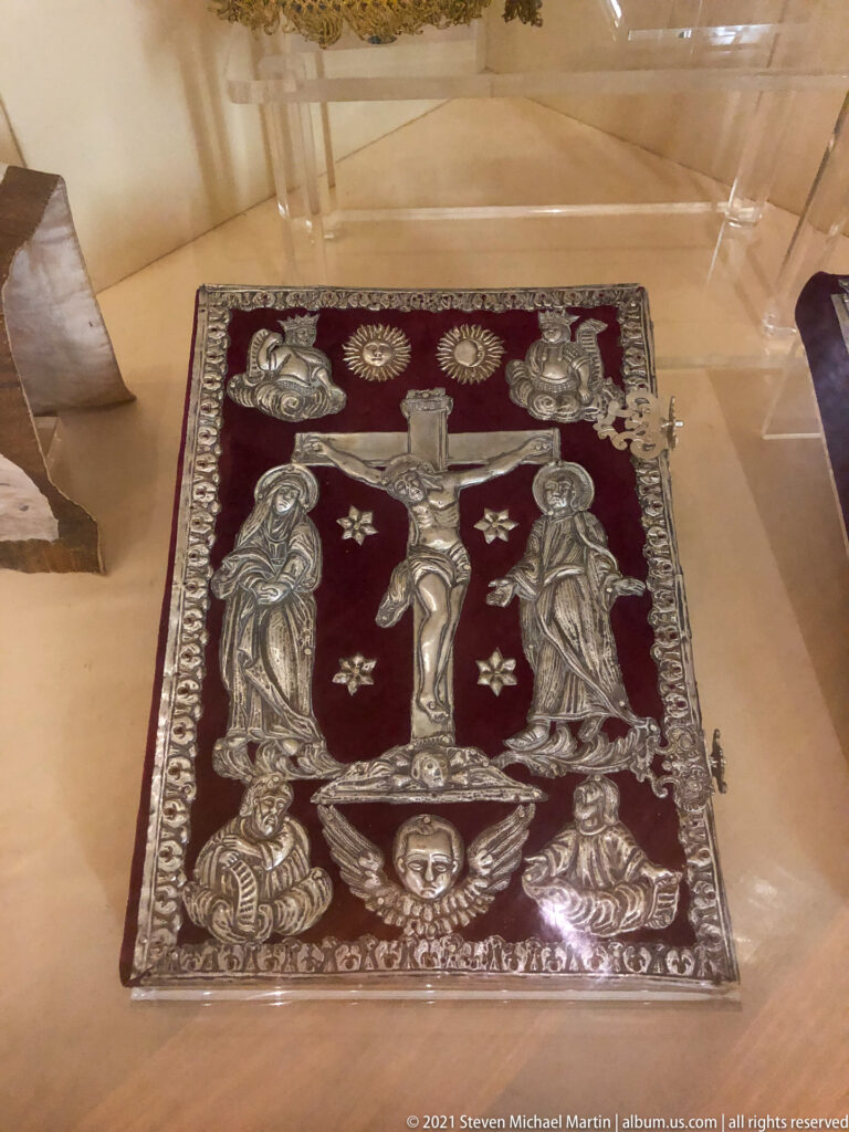 Rectangular red liturgical textile with embroidered Crucifixion, flanking figures, smaller motifs, and ornate border in a display case.