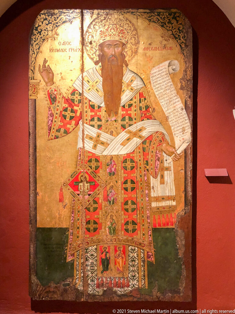 Full-length bearded bishop saint in richly patterned vestments, holding an inscribed scroll and raising one hand in blessing.
