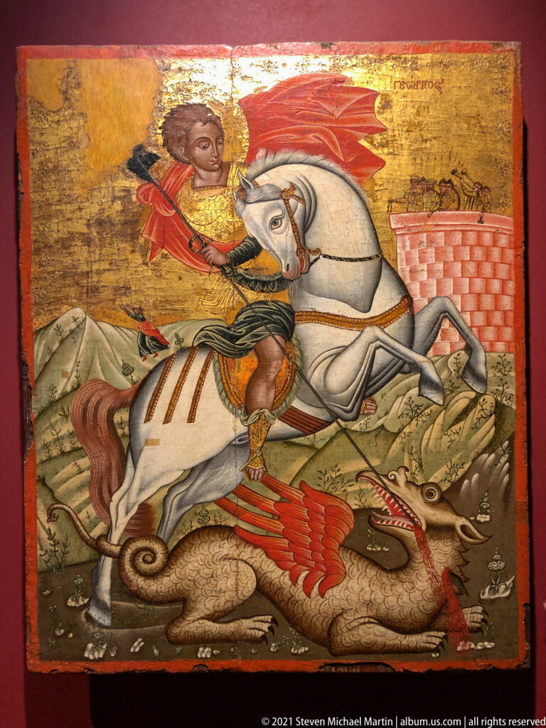 Saint George in a red cloak rides a white horse over a dragon beside a crenellated wall.