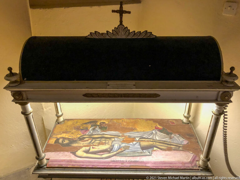 Painted icon displayed horizontally beneath a dark arched canopy supported by columns and topped with a cross.