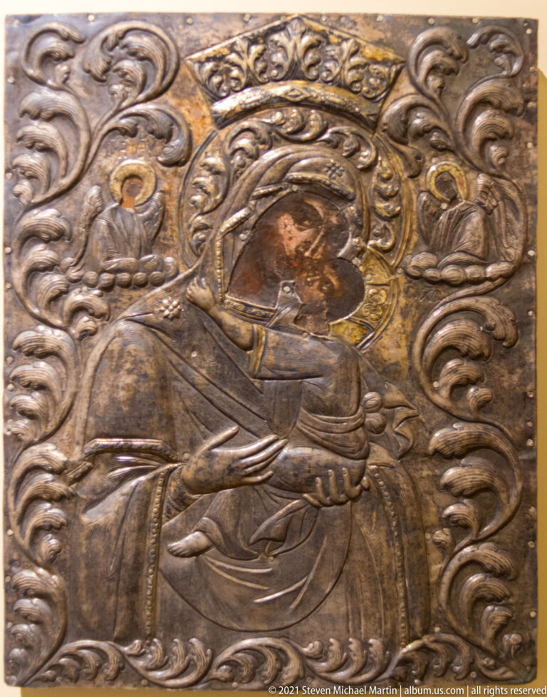 Embossed silver-toned icon covering with seated Virgin and Child amid dense scrolling relief ornament.