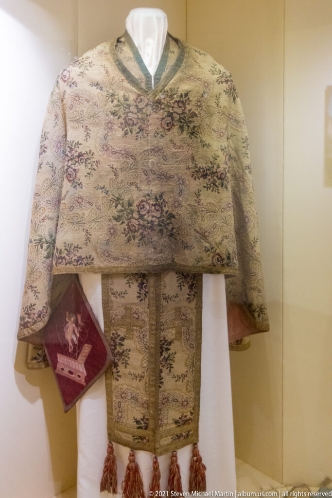 Pale embroidered Orthodox liturgical vestments on a mannequin, with floral patterns, decorative bands, tassels, and red textile detail.