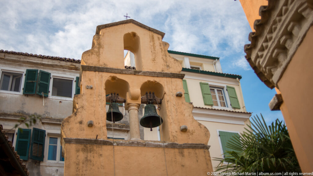 Weathered plaster bell gable with several bells and arched openings between neighboring buildings in Corfu.