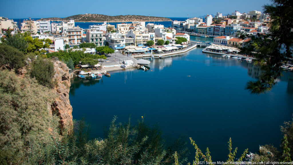 Lake Voulismeni seen from above, with boats, waterfront buildings, cafés, and rocky vegetation around the blue basin.