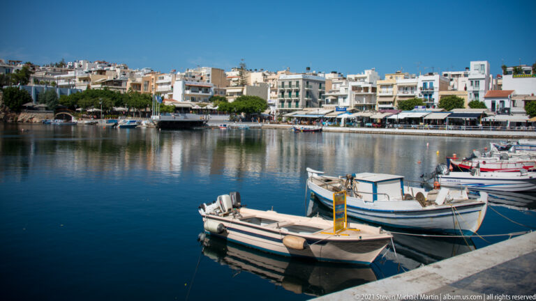 Small white fishing and leisure boats moored on calm blue water, with Agios Nikolaos waterfront buildings across the harbor.