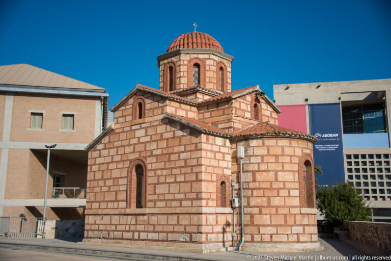 Small domed Orthodox church with alternating red brick and pale stone bands, arched windows, tiled roofs, and octagonal drum.