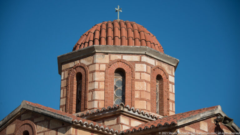 Close view of an octagonal church drum with red-brick arches, pale stone bands, terracotta dome tiles, and a metal cross.