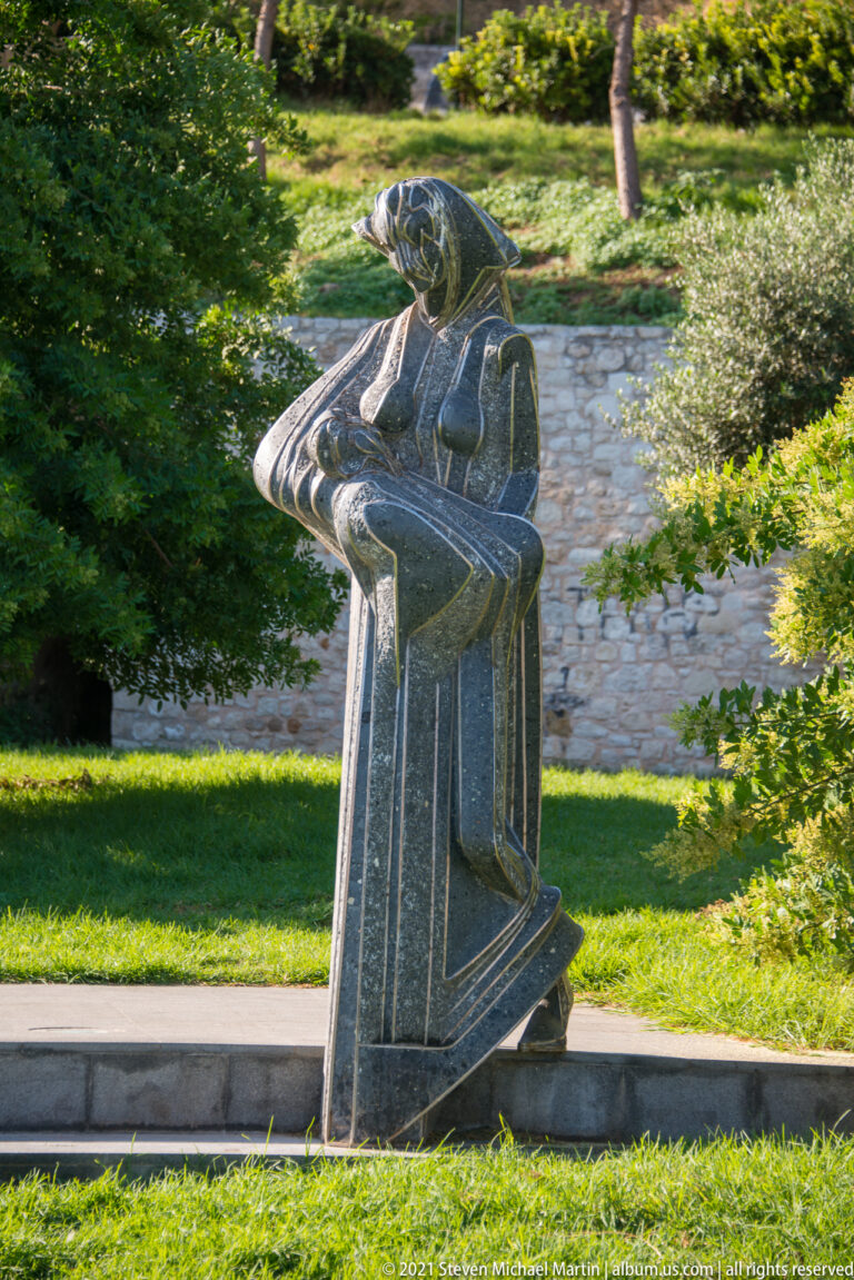 Dark stone abstract sculpture of a seated human figure beside a path in a green landscaped park.