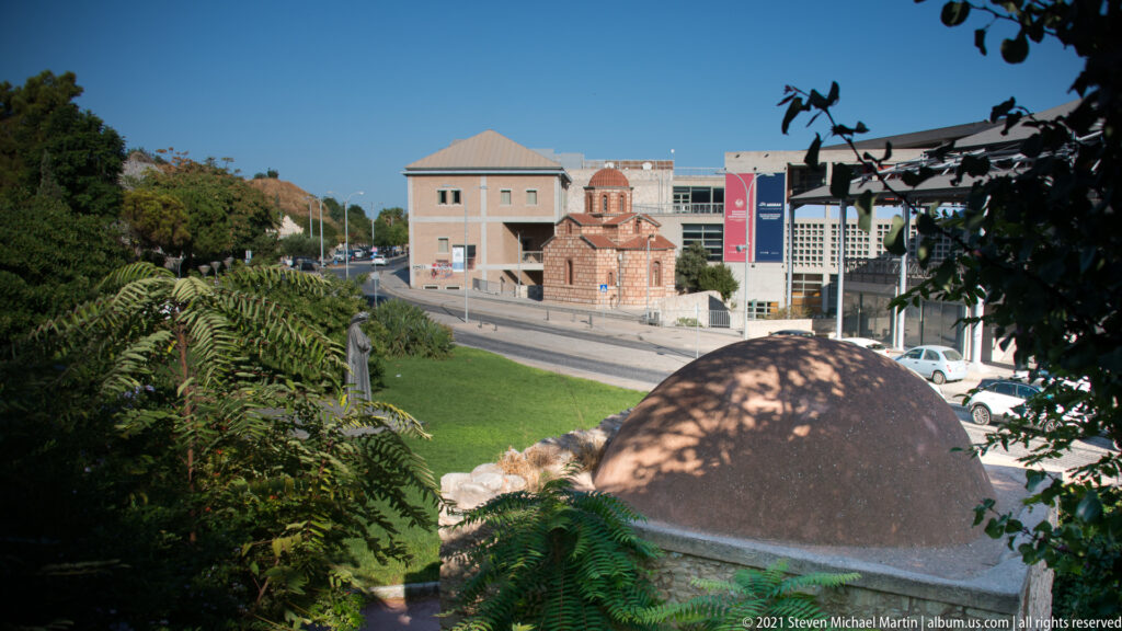 Terracotta dome in foreground with a small brick-and-stone church and modern institutional buildings across landscaped grounds.