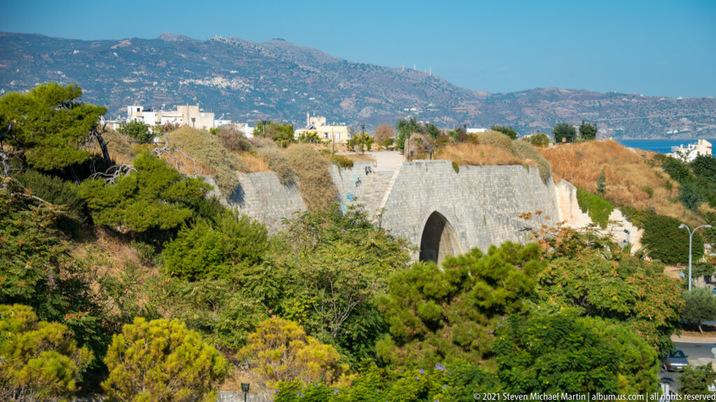 Elevated view of Heraklion's Venetian walls and arched passage amid dense trees, with mountains and city buildings beyond.