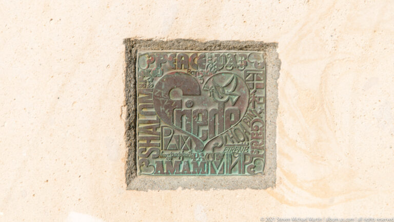 Square decorative relief mounted on a pale wall with stylized Heraklion lettering and multilingual words in green and reddish tones.
