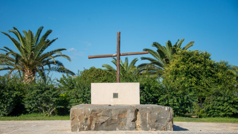 Simple wooden cross mounted on a white pedestal and dark stone base in a landscaped area with palm trees.