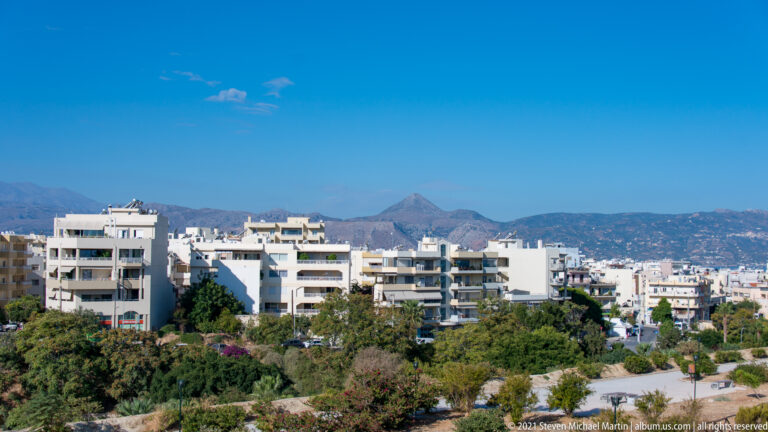 White apartment buildings above dense greenery in Heraklion, with dry rugged mountains stretching across the distant horizon.