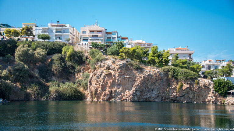 Rocky shrub-covered bluff rising from Lake Voulismeni, topped by white apartment and hotel buildings under a blue sky.