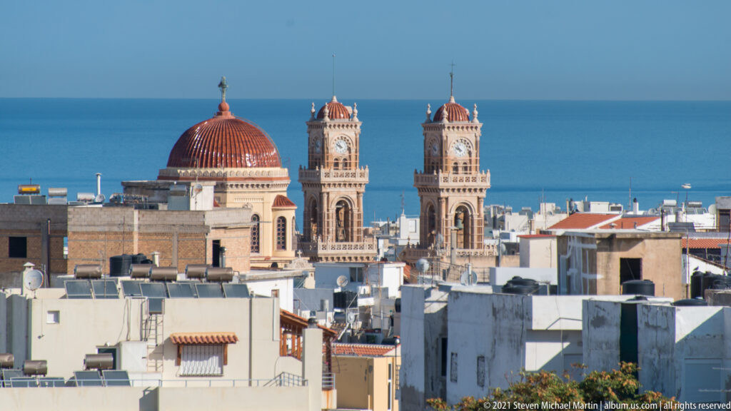 Saint Minas Cathedral rising above Heraklion rooftops with a red central dome, twin bell towers, and blue sea behind.
