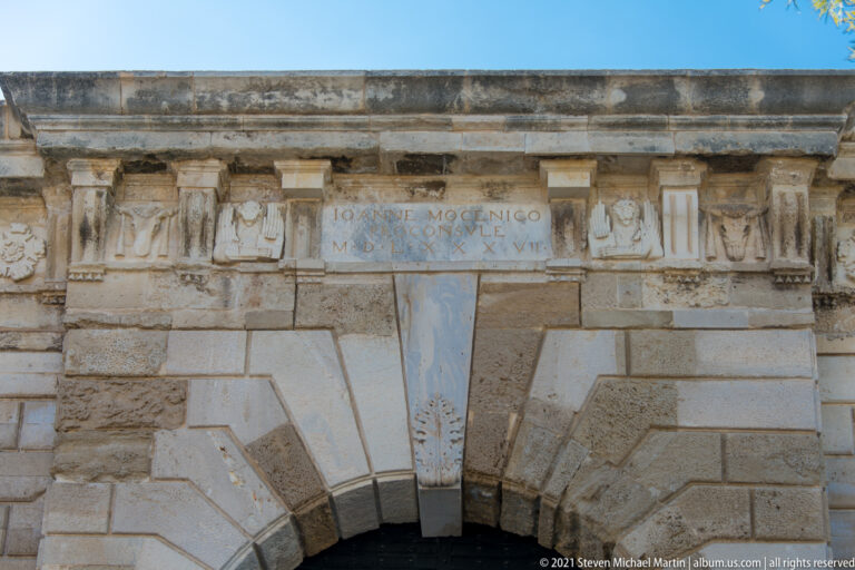 Close view of a Venetian stone gate with Latin inscription, carved relief panels, and radiating rusticated voussoirs above the arch.