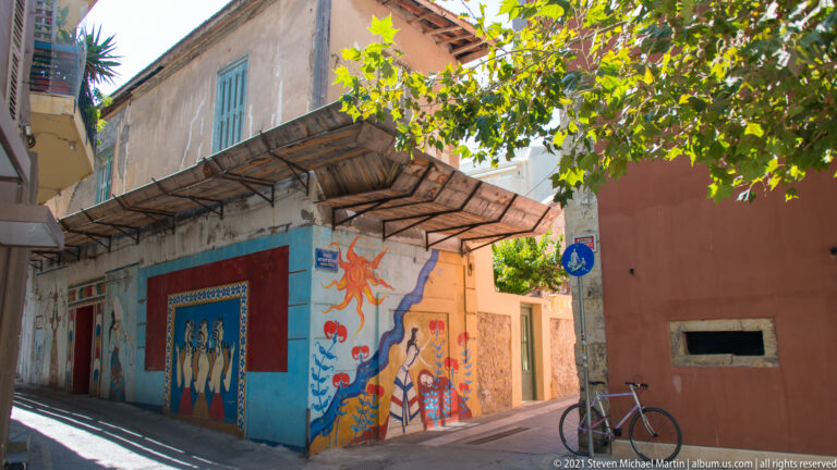 Colorful murals on a low building along a narrow Heraklion street, with a bicycle and leafy tree casting shade.