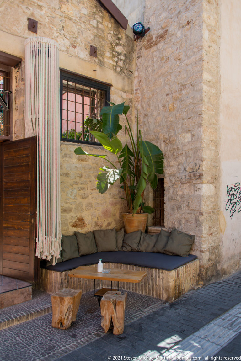 Stone-walled outdoor seating alcove with dark cushions, potted tropical plants, wooden shutter, and low rustic table.