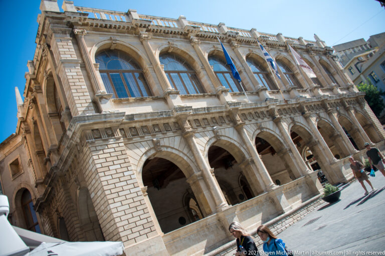Oblique daylight view of Heraklion's Venetian Loggia with ground-floor arcades, tall upper arches, rusticated stonework, and flags.