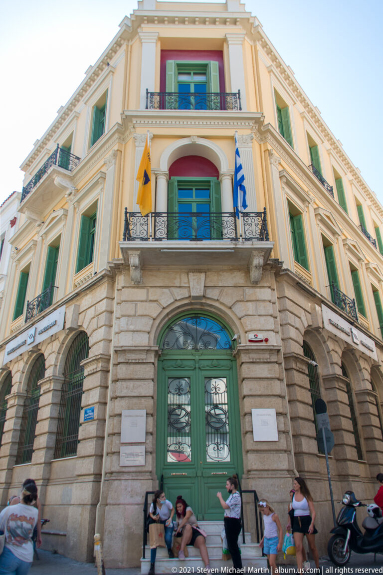 Pastel civic building with green shutters, iron balconies, arched green entrance, and Greek, EU, and yellow flags.