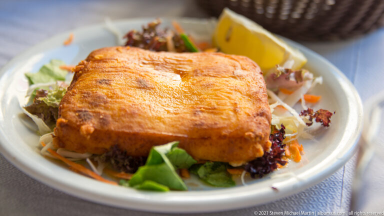 Golden-brown rectangular baked cheese on mixed greens with a lemon wedge on a white plate.