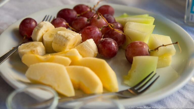 Plate of mixed fresh fruit with red grapes, banana slices, pale melon, and peeled yellow fruit, with forks at the edge.