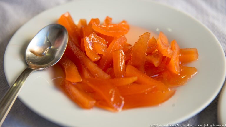 Glossy orange strips of preserved citrus peel in syrup on a small white plate with a teaspoon.