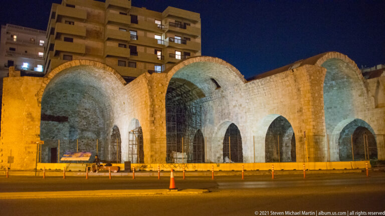 Illuminated stone-vaulted Venetian arsenal bays beside a road in Heraklion, with modern apartment buildings rising behind.