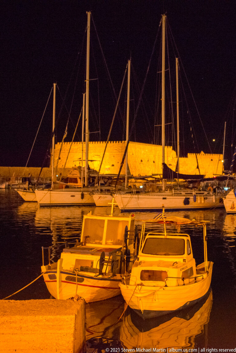 Small boats and yachts moored at night in Heraklion's Venetian harbor, with an amber-lit stone harbor wall beyond.
