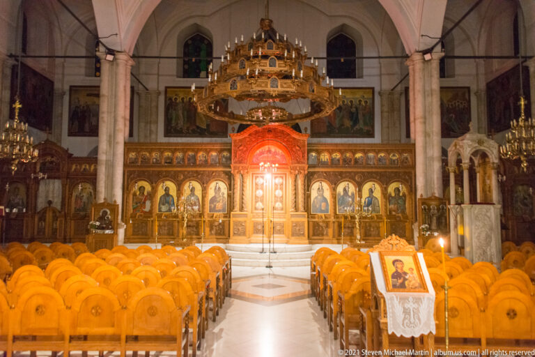 Central aisle of Saint Titus Church with rows of wooden chairs, gilded iconostasis, icons, chandelier, and pale stone arches.