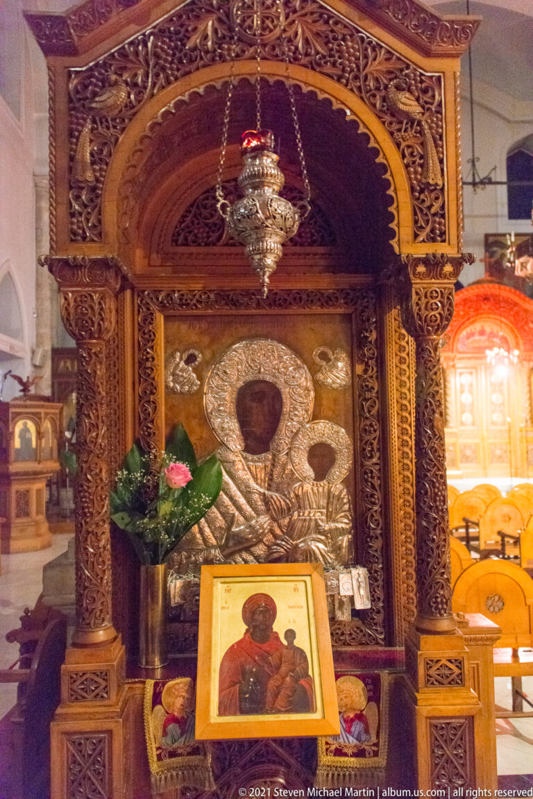 Ornate carved wooden icon shrine with twisted columns, hanging lamp, flowers, silver-toned backing, and framed icon.