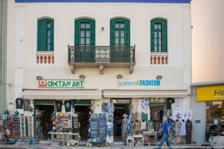 Souvenir and clothing shops below a pale façade with green shuttered windows and a wrought-iron balcony in Agios Nikolaos.