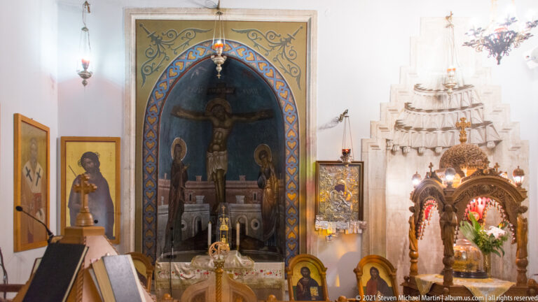 Side chapel with painted Crucifixion in a blue arched recess, altar vessels, icons, lamps, candles, and carved shrine.