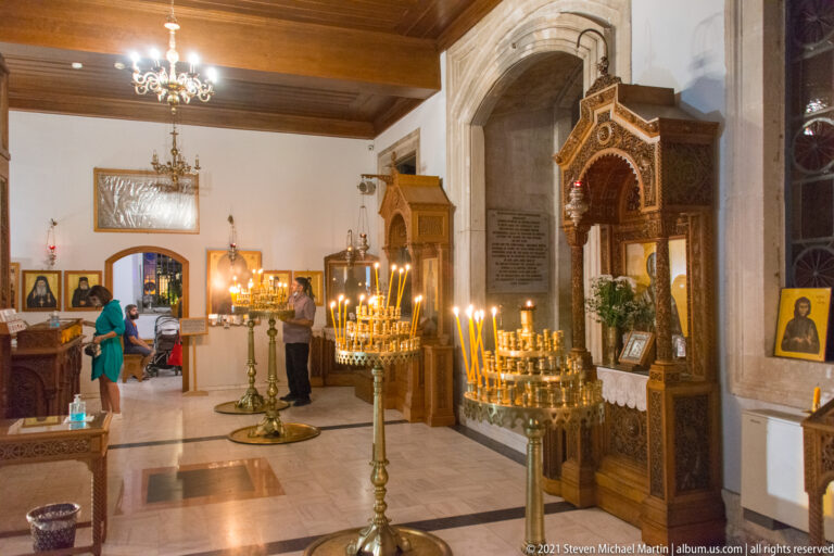 Brass candle stands filled with burning taper candles beside an ornate wooden shrine, with visitors and church interior beyond.